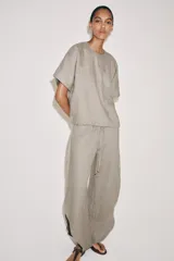 Top de lino de color beige, con cuello redondo, manga corta, bolsillos frontales aplicados y bajo ajustable con cordones. Cuenta con abertura en la espalda y cierre de botón.