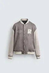 Cazadora estilo varsity o bomber de hombre, color gris oscuro con efecto ante, mangas en tono gris claro y puños, cuello y bajo con rib elástico a rayas blancas y grises. Presenta un parche con la letra 'R' en el pecho derecho y un bordado con texto en el pecho izquierdo.