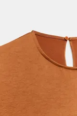 Camisa fluida color tostado de manga corta con vuelo y bajo abullonado con elástico. Cuenta con cuello redondo y cierre en la espalda con abertura y botón.