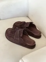 Zueco tipo mocasín de gamuza color marrón chocolate, con detalle de costuras visibles en el borde y cordones decorativos con borlas en el empeine.
