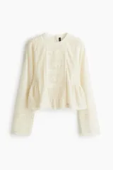 Blusa de manga larga confeccionada en malla plumeti color crema, con detalles de encaje, bordados y alforzas. Presenta cuello redondo, cierre con botón en la nuca y un corte con frunces laterales en la cintura que aportan volumen al bajo.