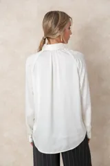 Blusa de satén color marfil con brillo sutil, cuello clásico, cierre frontal con botones y mangas largas con puños abotonados. Presenta un corte fluido y elegante.