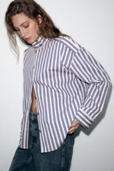 Camisa de manga larga con cuello subido y trabillas en los hombros. Presenta un diseño de rayas verticales en tonos azul, beige y marrón, con cierre frontal de botones.