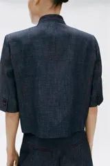 Blazer de manga corta confeccionado en lino, color azul marino, con cuello alto, cierre cruzado con un botón y pespuntes en contraste de color rojo. Cuenta con hombreras y bolsillos delanteros ribeteados.