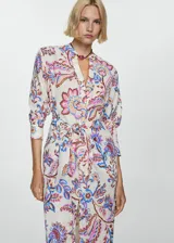 Vestido midi de algodón con estampado floral multicolor sobre fondo crudo. Presenta escote en pico, mangas largas con puños abotonados y un lazo decorativo en la cintura que crea un efecto drapeado, con una abertura frontal.