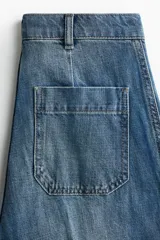 Pantalón de denim azul con lavado medio, corte de pierna recta y tiro alto. Presenta bolsillos de parche delanteros con solapa, trabillas para cinto y detalles de pliegues marcados en el centro de las piernas.