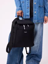 Mochila negra de estilo urbano con solapa superior, cierre con cordón ajustable y bolsillo frontal con etiqueta de la marca Rusty Supply. Cuenta con correas ajustables y acolchadas.