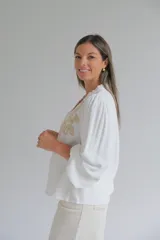 Blusa blanca de corte holgado con cuello mao y cierre frontal de botones. Presenta mangas largas abullonadas con puños elásticos y un delicado bordado en color beige en la parte superior delantera.