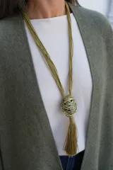 Collar largo de estilo artesanal con cordones de hilo color verde oliva, que presenta un dije central esférico compuesto por hilos metálicos entrelazados y una borla colgante del mismo tono.