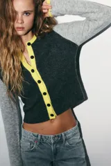 Chaqueta de punto con diseño color block, cuello redondo y manga larga. Presenta cuerpo en color gris oscuro, mangas en gris claro y detalles de ribetes en color amarillo vibrante. Cierre frontal con botones oscuros.