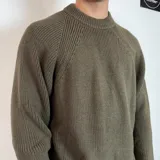 Sweater de tejido de punto grueso color verde militar, con cuello redondo y mangas largas tipo raglán. Presenta un corte holgado y una pequeña etiqueta con logo en el puño.