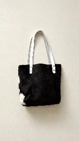Cartera tipo tote bag confeccionada artesanalmente en cuero con pelo, con diseño bicolor en negro y blanco. Presenta correas de hombro en cuero liso de color blanco sujetas con remaches metálicos.