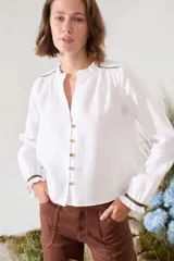 Blusa de lino color marrón con escote en V, cuello con volados y mangas largas con puños fruncidos que presentan detalles en color crudo, al igual que en los hombros. Cuenta con cierre frontal de botones.