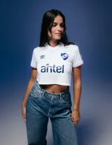 Camiseta de fútbol Umbro del Club Nacional de Football, modelo Home 2026 para mujer. Diseño blanco tipo crop con cuello polo, detalles en azul y rojo en cuello y mangas, logo de Umbro, escudo del club y sponsor Antel en el frente.