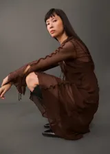 Vestido largo de gasa color marrón, con diseño evasé, cuello camisero y cierre frontal con lazos. Presenta mangas largas y detalles de volados en los hombros.