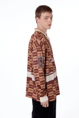 Camiseta oversize marrón con estampado de logos y texto "Blkmba" en toda la prenda. Tiene cuello en V con cuello tejido, escudo Mamba soccer en la manga y logo blkmba en el centro.