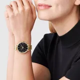 Reloj analógico Michael Kors con caja y malla de acero inoxidable en tono dorado. La esfera es de color negro con el logo de la marca grabado en relieve y cuenta con un bisel decorado con cristales brillantes.