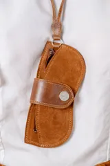 Estuche para lentes con correa de cuero vacuno, diseñado para colgar. Presenta un diseño abierto en gamuza color óxido con una banda frontal de cuero liso y cierre con botón metálico a presión.