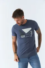 Remera de hombre color azul, con cuello redondo y manga corta. Presenta un diseño gráfico estampado en el frente con formas geométricas y letras en tonos blanco y gris.