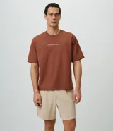 Remera de corte boxy y amplio, color marrón, con cuello redondo, mangas cortas y estampa frontal minimalista con texto en blanco.