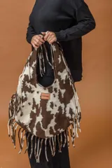 Bolso tipo hobo de gran capacidad con estampado animal print de vaca en tonos marrón y blanco. Presenta una etiqueta de cuero con el logo de la marca en el frente y un diseño de caída relajada.