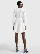 Vestido camisero corto color blanco, confeccionado en algodón orgánico. Presenta cuello en V, cierre frontal con botones, mangas largas abullonadas, cinturón de lazo ajustable en la cintura, bolsillos laterales y detalle de encaje calado en forma de escalera.