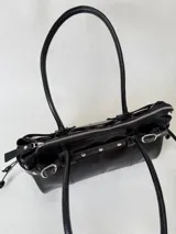 Cartera de cuero de tamaño medio con estructura rígida. Presenta una tira decorativa con tachas y hebillas metálicas en los extremos, además de lazos laterales. Cuenta con doble asa para llevar al hombro y cierre superior.