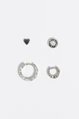 Set de cuatro pendientes de metal plateado que incluye un pendiente en forma de corazón, un pendiente circular con cristal central, una argolla mediana y una argolla pequeña, ambas con detalle de letras grabadas.