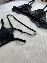 Conjunto de lencería de cuatro piezas en encaje negro. Incluye bralette con breteles regulables, tanga con tiras dobles ajustables en la cintura, portaligas con enganches desmontables y ligas para las piernas.
