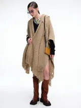 Ruana tejida de color beige con capucha integrada y terminaciones con flecos en el ruedo. Presenta un diseño envolvente y holgado, confeccionada en una mezcla de lana merino extrafina y viscosa.