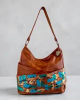 Cartera tipo hobo de cuero natural color marrón con arte pintado a mano en tonos turquesa y dorado en la base, de diseño envolvente y caída suave. Cuenta con correa de hombro y detalle de dije metálico colgante.