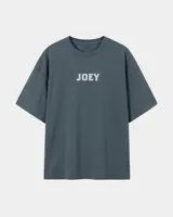 Remera oversize gris oscuro con corte holgado, mangas cortas y estampa trasera en blanco y negro con forma de estampilla que muestra a una persona con un gato y el texto 'Kaboa Industries'.