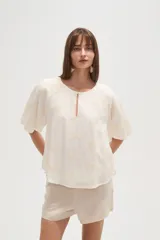 Blusa blanca de manga corta con escote redondo y abertura frontal con botón. Presenta bordados florales en tono beige distribuidos por toda la prenda.