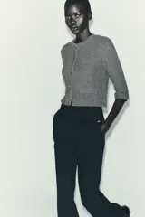 Chaqueta de punto grueso color gris, de cuello redondo y manga tres cuartos. Cierre frontal con botones joya dorados con detalles de pedrería. Cuenta con dos bolsillos frontales tipo parche.