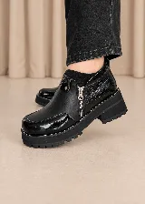 Zapato de cuero color negro con plataforma y suela dentada. Presenta una combinación de texturas que incluye detalles en charol y un sector con textura reptilada, además de cierre lateral y costuras en contraste.
