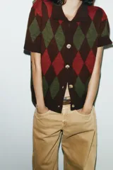 Chaqueta de punto de manga corta con cuello polo y cierre frontal de botones. Presenta un patrón de rombos (argyle) en tonos marrón oscuro, verde oliva y rojo.