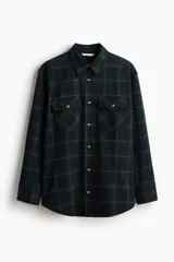 Camisa oversized de franela suave con estampado a cuadros en tonos azul marino, verde oscuro y beige. Presenta cuello, botonadura frontal con botones dorados, canesú en la espalda, hombros caídos, bolsillos superiores con solapa y botón dorado, y puños abotonados. El bajo es ligeramente redondeado.