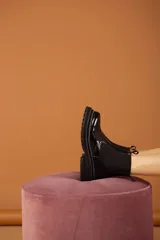 Botas tipo Chelsea de caña baja, confeccionadas en cuero charolado negro con acabado brillante. Presentan paneles laterales elásticos, suela track robusta con plataforma dentada y costuras visibles en el borde de la suela.