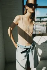 Top corsetero de tul semitransparente color beige, con escote cuadrado, tirantes anchos y cierre frontal con ganchos metálicos. Presenta bajo asimétrico y costuras reforzadas con ribetes a tono.