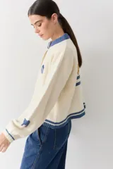 Cardigan de lana color crema con cuello redondo, cierre frontal de botones y mangas largas con puños ajustados. Presenta bordados decorativos en el frente que incluyen un sol, olas y estrellas, con detalles en azul en el cuello y el ruedo.