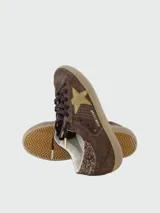 Zapatillas Golden Goose modelo Super Star, confeccionadas en gamuza color marrón con estrella lateral en gamuza dorada y detalle de glitter en el talón. Presentan cordones oscuros y suela de goma con efecto desgastado.