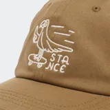 Gorro de béisbol color verde oliva con bordado frontal que incluye una flor sonriente con piernas y las letras "STANCE" rodeándola.