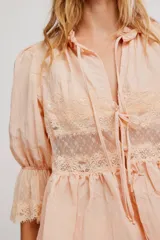 Camiseta de tela liviana color salmón, con terminaciones de encaje, escote en V profundo y botonadura al frente con tiras para anudar. Mangas tres cuartos abullonadas con puño elastizado y ruedo acampanado, cuello con volado.