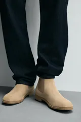 Bota estilo Chelsea de piel con acabado serraje color beige, con elásticos laterales para facilitar el calce, horma redonda y suela a tono con ligero tacón.