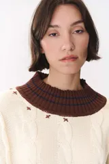 Buzo de tejido de punto con diseño de trenzas, color crema con detalles en marrón en cuello, puños y ruedo. Presenta líneas finas en contraste y pequeñas costuras decorativas en forma de cruz.