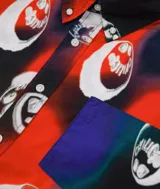 Camisa de manga corta con estampado multicolor de caras sonrientes sobre fondo rojo, azul y negro. Tiene cuello abotonado, bolsillo en el pecho y cierre frontal con botones.