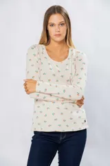 Remera de manga larga color crudo con estampado de flores pequeñas en tonos naranja y verde. Tiene cuello redondo y pin metálico con logo personalizado en el ruedo.