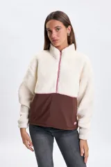 Buzo de cuello alto con medio cierre frontal, confeccionado en una combinación de tejido tipo sherpa en la parte superior y nylon en la parte inferior. Presenta un diseño bicolor en tonos crudo y marrón, con detalles de cierre en color rosado.