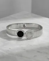 Esclava de acero inoxidable con diseño rígido y flexible, que presenta dos piezas circulares centrales: una con piedra de ónix negro y otra con nácar blanco, ambas rodeadas por un borde con números romanos grabados.