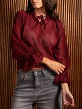 Blusa de plumeti color bordó, con cuello alto ajustable mediante lazo, mangas largas con puños elásticos y detalle de volados en las terminaciones.